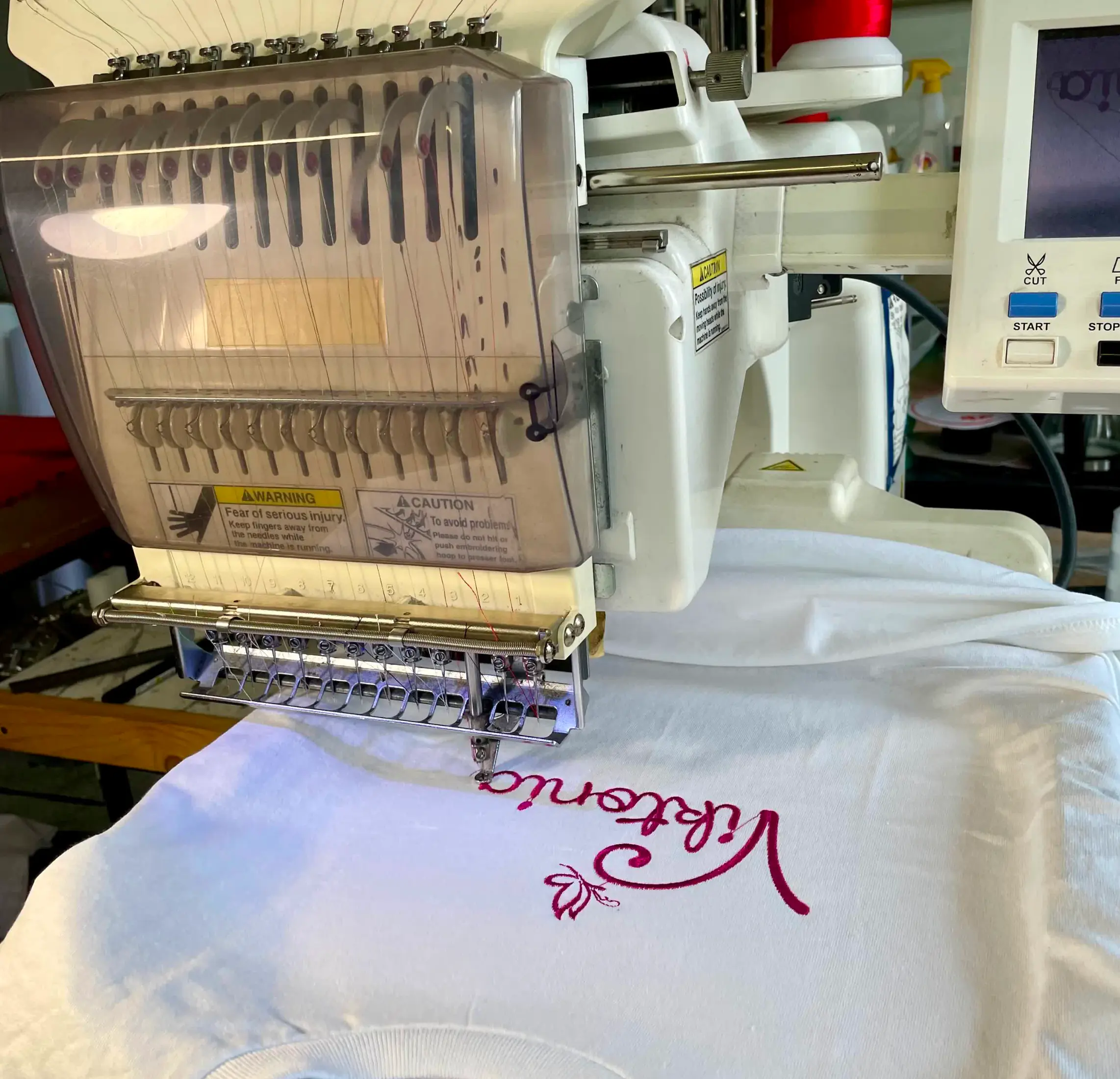 An embroidery machine is stitching a white fabric with the word "Welcome" in red thread.