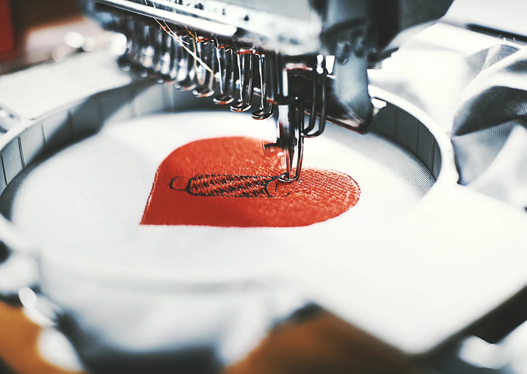 A sewing machine embroidering a red heart onto fabric in a circular embroidery hoop.