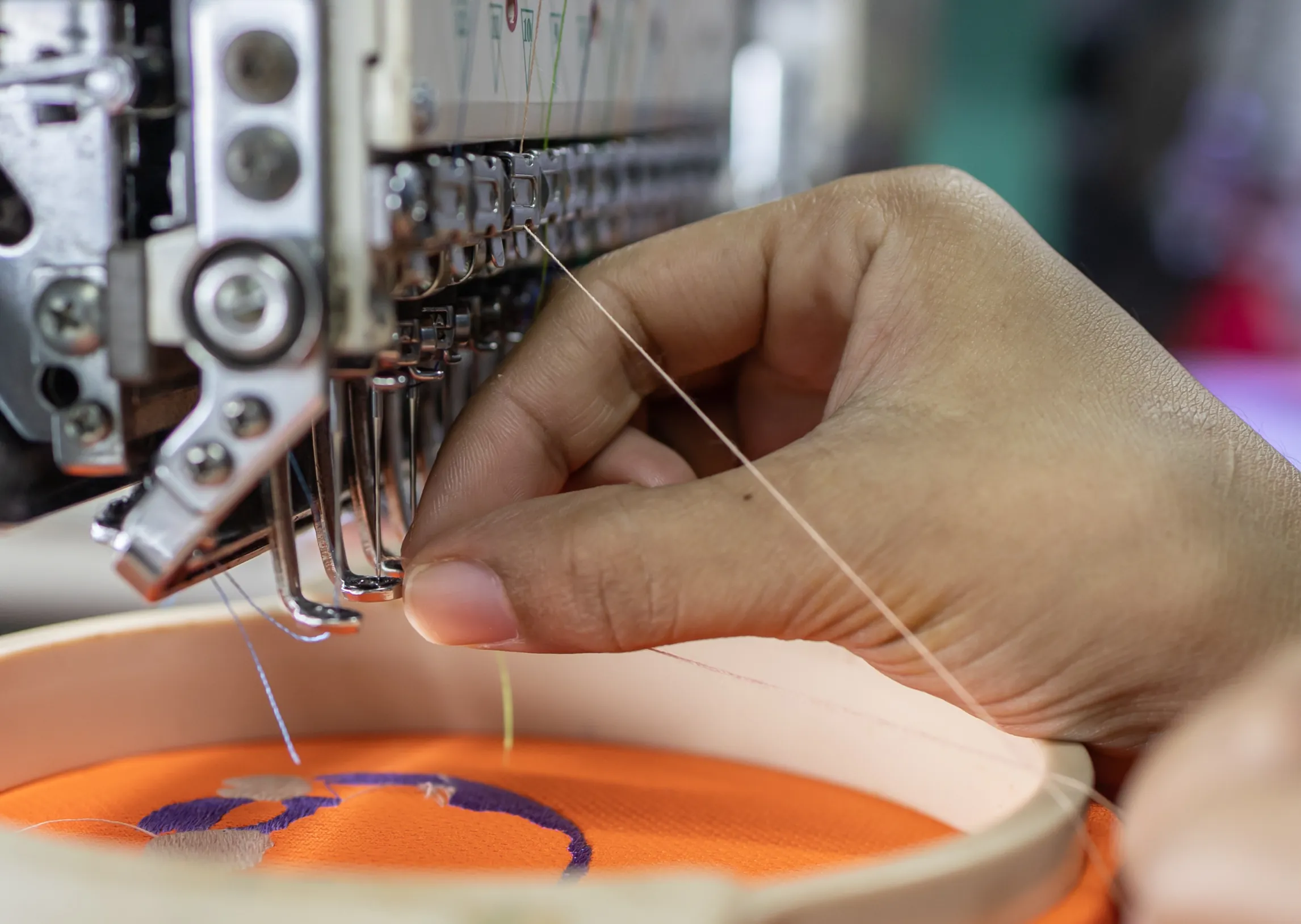 Close-up of a hand threading a needle on an embroidery machine, with an orange fabric in the hoop.